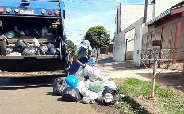 Fotografia mostra um coletor de lixo uniformizado recolhendo sacos plásticos em uma rua residencial atrás de um caminhão de coleta de lixo. Do lado, calçada e casas.