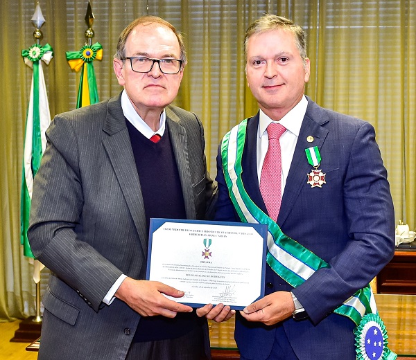 Fotografia de dois homens brancos lado a lado, vestidos de terno, e segurando em frente um diploma. À esquerda, o presidente do TRT-PR, desembargador Célio Waldraff. À direita, o ministro Douglas Rodrigues, com faixa da comenda nas cores verde e branco, e a medalha presa ao paletó.