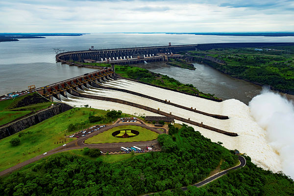 Foto aérea da usina da itaipu com o lago ao lado esquerdo e a barragem de concreto ao centro e do lado direito, o vertedouro com água.