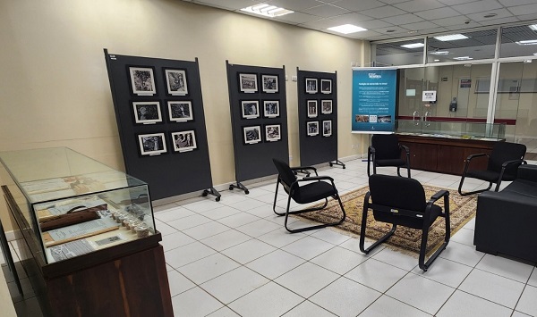 Fotografia de saça com cadeiras, um sofá, duas mesas com tampo de vidro com objetos expostos e, ao fundo, paineis com fotos expostas.