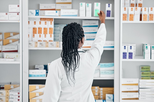 Mulher preta com cabelos com tranças, de costas, usa um jaleco branco e ergue o braço direito para pegar um produto em prateleira refleta de medicamentos.