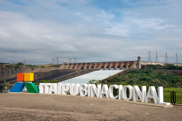Fotografia de uma letreiro da Itaipu Binacional com a usina ao fundo
