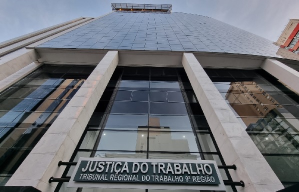 Fotografia da fachada em vidro do TRT-PR. A imagem mostra a vista a partir do chão para o alto do prédio. Destaca-se letreiro escrito: Justiça do Trabalho, tribunal regional do trabalho da 9ª Região