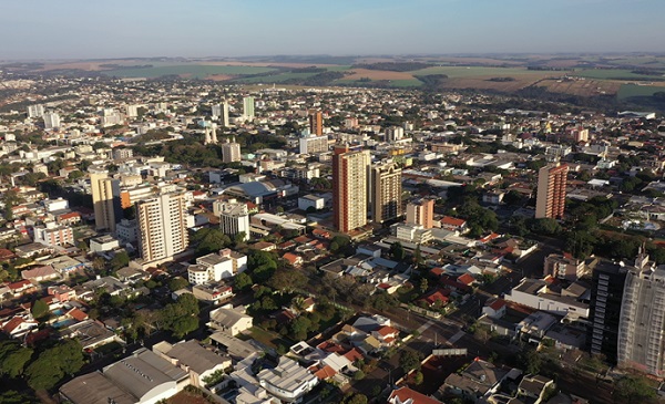 Imagem área de cidade com alguns prédios e várias casas entre árvores. Ao fundo, áreas de plantação agrícola.
