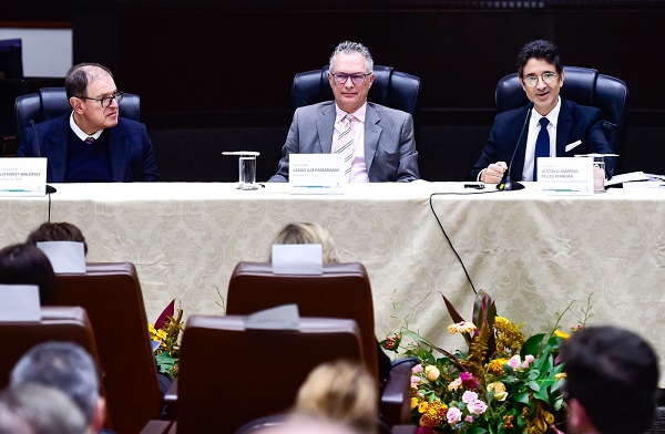 Fotografia de três homens sentados em frente a uma mesa. Todos vestem terno. O da esquerda fala ao microfone e olha para a plateia em frente.