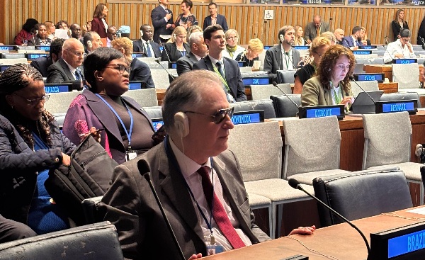 Fotografia do desembargador Ricardo Tadeu, homem branco vestido de terno e que utiliza óculos escuro, sentado na plenária da ONU com outras pessoas atrás dele.
