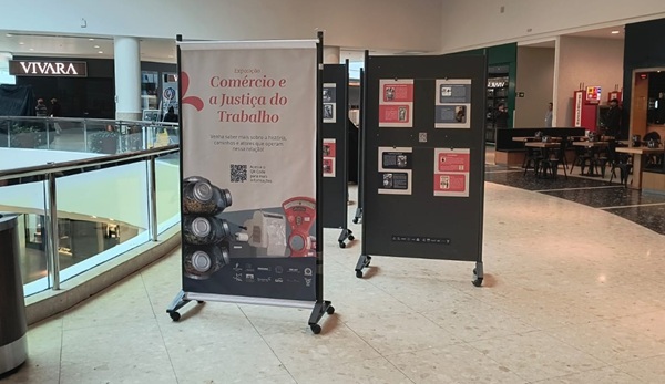 Fotografia de um ambiente de circulação com biombos, com destaque para um banner escrito Comércio e Justiça do Trabalho, e lojas ao fundo.