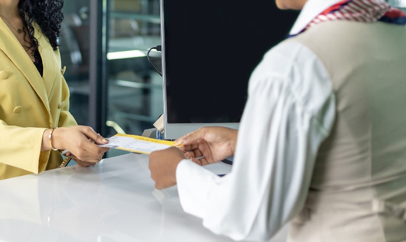 Fotografia de um balcão em que duas mulheres seguram um envelope, uma com uniforme de trabalho. O corpo de ambas aparece parcialmente na altura dos braços;.