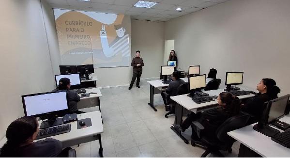 Fotografia de uma sala de aula com projeção na parede escrito Currículo para o primeiro emprego. Pessoa de pé em frente aos alunos, sentados em frente à computadores.
