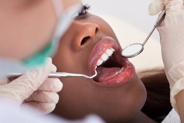 Fotografia de mulher preta sendo analisadas por dentista, que tem luvas e instrumentos odontológicos em mãos