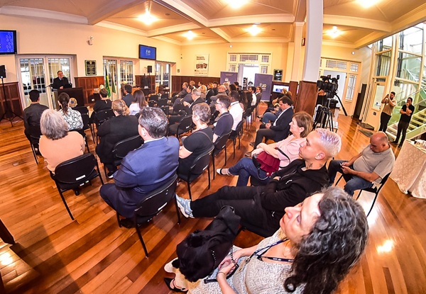 Fotografia de um grupo de pessoas de costas sentados em cadeiras no foyer do Tribunal com o presidente ao fundo falando em uma tribuna