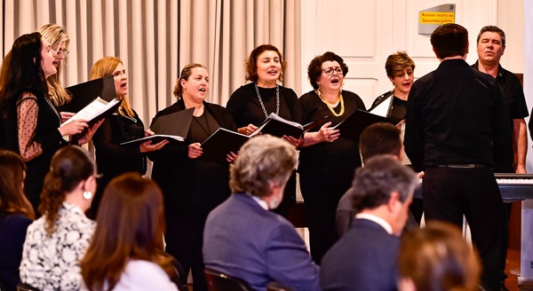 Fotografia de um grupo de cerca de dez pessoas de roupas pretas cantando em frente à plateia