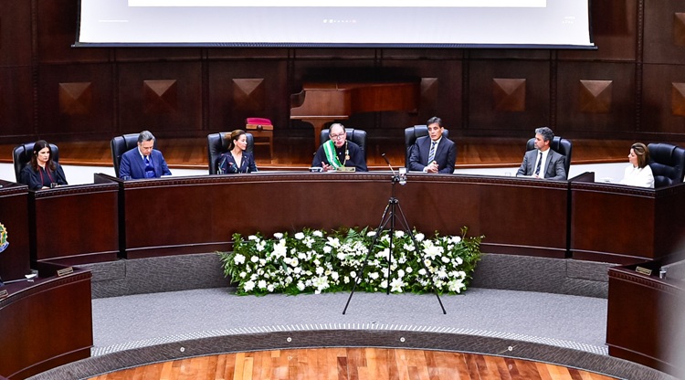 Fotografia da mesa diretiva da cerimônia com sete pessoas, três mulheres e quatro homens sentados em frente à balcões de madeira