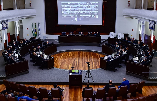 Fotografia do plenário, apartir das galerias superiores, mostra ao fundo um telão e em frente a mesa diretiva com as mesas dos desembargadores que formam o Pleno à direita e à esquerda. 