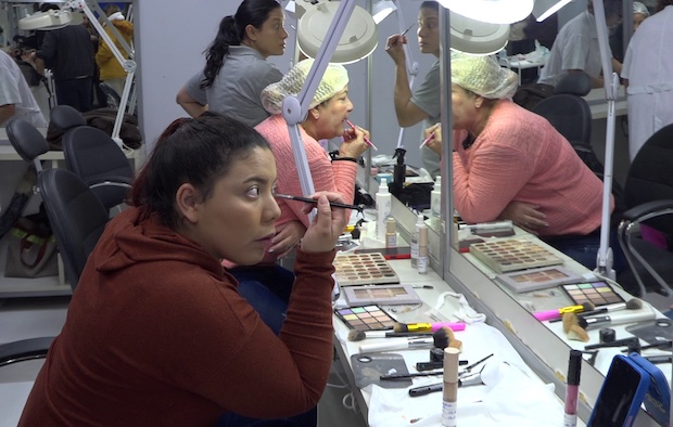 Mulheres se maqueiam em frente a espelhos com materiais de uso profissional
