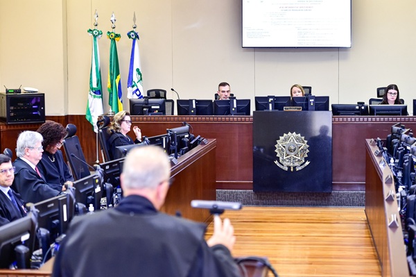 Fotografia da se&ccedil;&atilde;o especializada com a mesa diretiva ao fundo, com duas mulheres e um homem; no meio da imagem dois desembargadores e duas desembargadoras sentados em cadeiras em frente a telas de computadores e, em primeiro plano, um advogado de costas em p&eacute; faz sustenta&ccedil;&atilde;o oral.