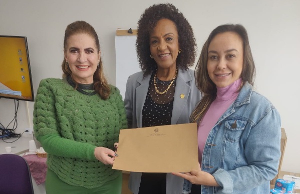 Fotografia de três mulheres em pé lado a lado. Elas seguram em conjunto um envolvepe ao centro da imagem. Todas sorriem