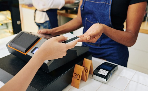 Cliente pagando a funcion&aacute;ria com cart&atilde;o de cr&eacute;dito em estabelecimento comercial. Ambos est&atilde;o de p&eacute; em frente a um balc&atilde;o com caixa registradora em frente.