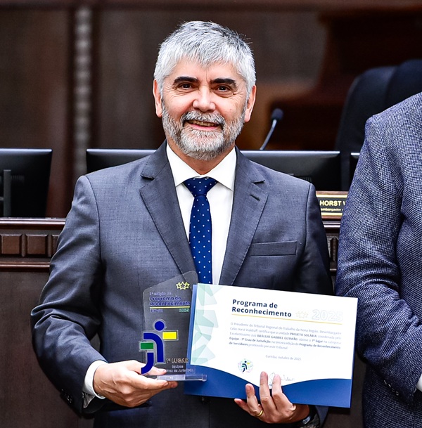 Fotografia do juiz Braulio Gusm&atilde;o, de terno cinza, camisa branca e gravata azul. ele segura um trof&eacute;u e certicado. Ele &eacute; homem branco, de barba e cabelos grisalhos.