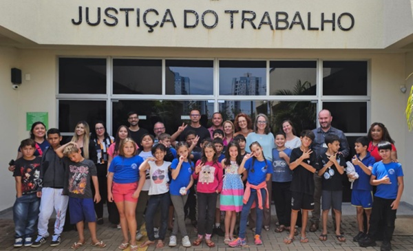 Fotografia de um grupo de crian&ccedil;as com adultos em frente &agrave; fachada do pr&eacute;dio da Justi&ccedil;a do Trabalho em Foz do Igua&ccedil;u.