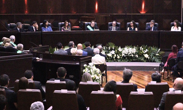 Forografia do plen&aacute;rio com plateia de costas no primeiro plano e, ao fundo, a mesa de autoridades da cerim&ocirc;nia, com o presidente Artio Mazurkevic discursando. H&aacute; nove autoridades sentadas em frente &agrave; mesa, cinco homens e quatro mulheres