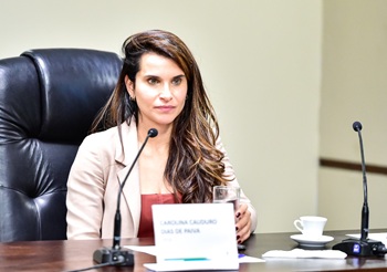 Fotografia de uma mulher branca de cabelos longos sentada em cadeira preta em frente a uma mesa com micrfone.