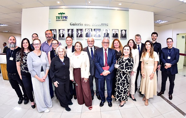 Fotografia de um grupo de cerca de 20 pessoas perfiladas lado a lado em duas linhas e atr&aacute;s um quadro com fotografias de diretores da EJ do TRT-PR
