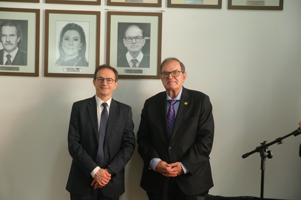 Fotografia de dois homens brancos de terno lado a lado em p&eacute;. Atr&aacute;s de ambos,uma galeria de fotografias