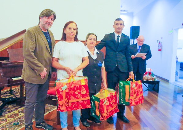 Fotografia de quatro pessoas perfiladas lado a lado, tr&ecirc;s delas com sacolas com cestas natalinas. 