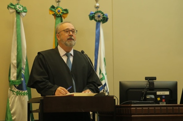 Fotografia de um homem grisalho de barba e &oacute;culos em p&eacute;, falando em frente a uma tribuna de madeira como microfone. Ao fundo, bandeiras do Paran&aacute;, Brasil e TRT
