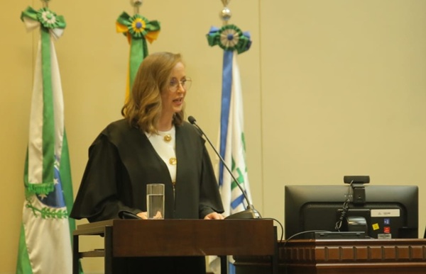 Fotografia de uma mulher loira em p&eacute;, falando em frente a uma tribuna de madeira como microfone. Ao fundo, bandeiras do Paran&aacute;, Brasil e TRT