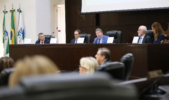Fotografia de uma bancada de madeira, com cinco pessoas sentadas atr&aacute;s com microfones em frente, quatro homens e uma mulher. Ao centro, o ministro Freire Pimenta discursa