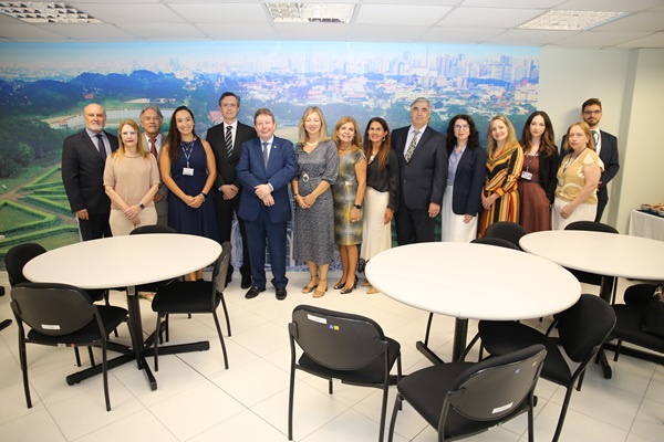 Grupo de 15 pessoas perfiladas lado a lado, sendo nove mulheres e seis homens. Em frente ao grupo, h&aacute; cadeiras e mesas