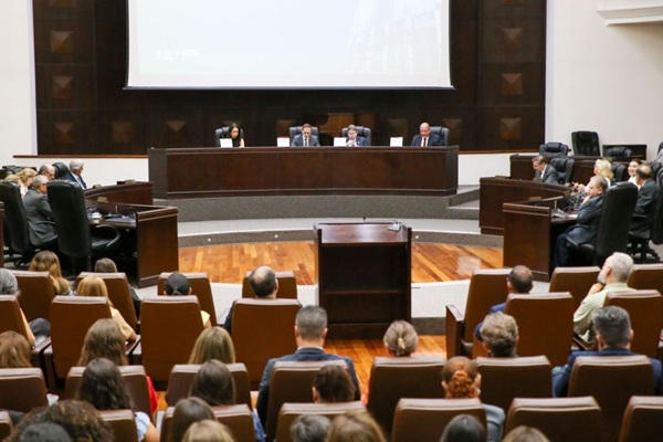 Fotografia de uma bancada de madeira com quatro pessoas sentadas atr&aacute;s em frente &agrave; microfones, sendo tr&ecirc;s homens e uma mulher. Em primeiro plano, diversas pessoas na plateia de costas