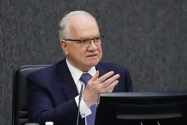 Fotografia de homem sentado em cadeira de couro em frente a um computador e microfone. Ele tem cabelo e bigode grisalho e &oacute;culos e veste terno com gravata azul.