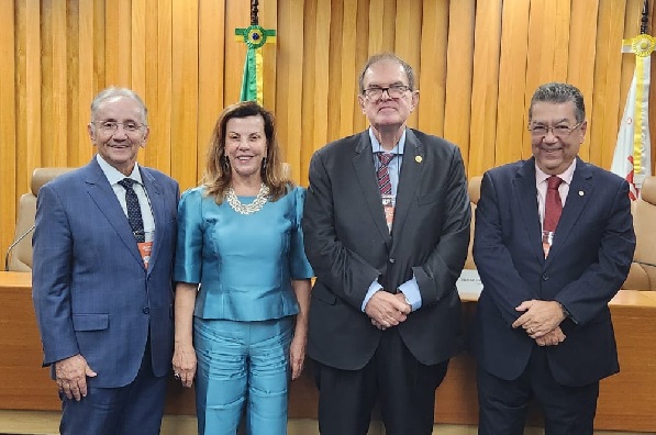Fotografia de quatro pessoas em p&eacute;, lado a lado, sendo tr&ecirc;s homens e uma mulher. atr&aacute;s deles, uma mesa de madeira e duas bandeiras, uma do Brasil e outra do TST