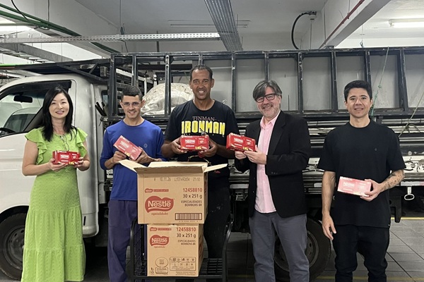 Fotografia de cinco pessoas lado a lado em frente a um caminh&atilde;o pequeno, sendo uma mulher e quatro homens. Cada pessoa segura uma caixa de bombom. em frente ao grupo, duas caixas de papel&atilde;o uma sobre a outra