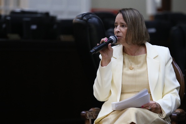 Fotografia de mulher sentada em cadeira de madeira com microfone na m&atilde;o esquerda. Ela tem cabelo na altura do ombro e usa blazer e vestido cor creme.