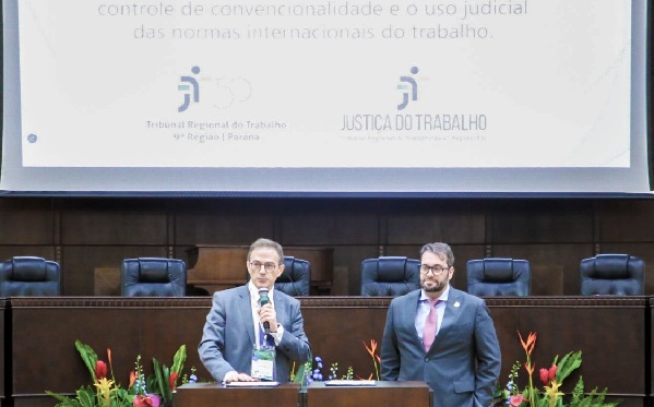 Fotografia de dois homens em p&eacute; vestindo ternos em frente a uma mesa de madeira. O da esquerda segura um microfone. Logo atr&aacute;s, arranjos de flores e ao fundo tela escrita controle de convencionalidade e o uso judicial das normas internacionais do trabalho