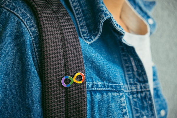 Fotografia aproximada de uma pessoa de jaqueta jean usando mochila. Imagem mostra apenas a &aacute;rea peitoral. Preso na al&ccedil;a da mochila, um broch&ecirc; com o s&iacute;mbolo do infinito colorido, s&iacute;mbolo de neurodiversidade e autismo