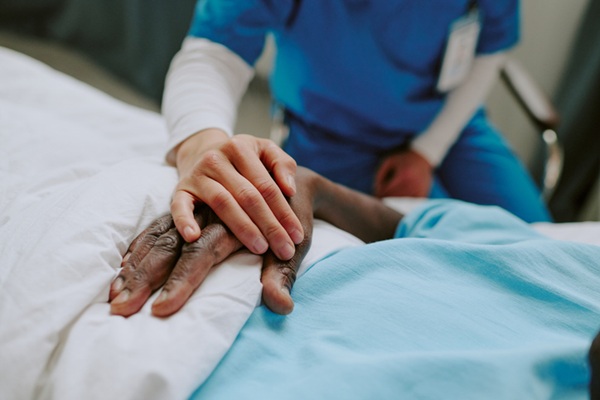 A imagem mostra uma cena de acolhimento em ambiente hospitalar. Uma pessoa vestindo um uniforme azul (possivelmente um profissional de sa&uacute;de) segura suavemente a m&atilde;o de um paciente sobre um len&ccedil;ol branco. A pele do paciente &eacute; negra, contrastando com a m&atilde;o de pele clara do profissional. O foco est&aacute; no gesto de cuidado e empatia. A imagem transmite humaniza&ccedil;&atilde;o no atendimento m&eacute;dico.
