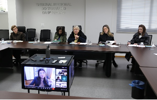 Foto de uma reuni&atilde;o em sala do Tribunal Regional do Trabalho da 9&ordf; Regi&atilde;o. Cinco mulheres est&atilde;o sentadas &agrave; mesa, em reuni&atilde;o presencial. &Agrave; frente, um monitor exibe uma videoconfer&ecirc;ncia com outras participantes. O ambiente &eacute; formal, com m&oacute;veis escuros e ilumina&ccedil;&atilde;o interna. As pessoas est&atilde;o focadas na discuss&atilde;o, utilizando materiais de apoio sobre a mesa.