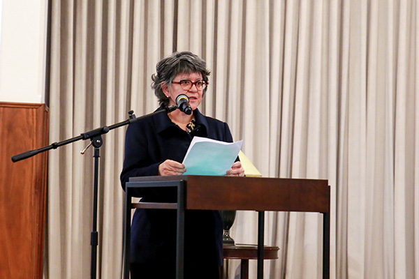 A imagem "aposentados 3 interna.jpg.jpeg" mostra uma mulher proferindo um discurso em um ambiente formal ou institucional.  Aqui est&atilde;o os principais detalhes da imagem:  Elemento Central: Uma mulher de pele clara, com cabelos curtos e grisalhos, usando &oacute;culos de arma&ccedil;&atilde;o escura e grossa. Ela veste um casaco escuro (aparentemente preto ou azul-marinho) estruturado e fechado.  A&ccedil;&atilde;o: Ela est&aacute; em p&eacute; atr&aacute;s de um p&uacute;lpito de madeira com estrutura met&aacute;lica escura, segurando algumas folhas de papel branco com as duas m&atilde;os, sugerindo que est&aacute; lendo um discurso ou notas. Ela fala diretamente ao microfone, que est&aacute; posicionado &agrave; sua frente em um pedestal articulado.  Plano de Fundo: O fundo &eacute; composto por uma cortina de tecido claro (bege ou off-white) com pregas verticais bem marcadas, que ocupa a maior parte da cena. &Agrave; esquerda, h&aacute; uma estrutura de madeira vertical, possivelmente parte de um painel ou coluna. Atr&aacute;s do p&uacute;lpito, vislumbra-se parcialmente o topo de um abajur ou objeto decorativo sobre uma mesa pequena.  A ilumina&ccedil;&atilde;o &eacute; focada na palestrante, conferindo um tom formal e s&oacute;brio &agrave; situa&ccedil;&atilde;o.
