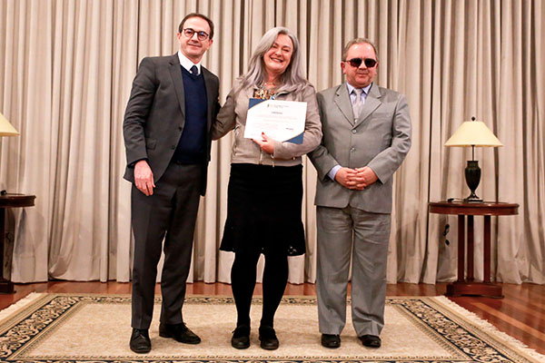A imagem retrata os desembargadores Arion Mazurkevic, Edmilson Antonio de Lima e a servidora aposentada Marlene Alessi. Eles est&atilde;o posando para a foto. Marlene segura o diploma com as duas m&atilde;os. Os tr&ecirc;s sorriem. 