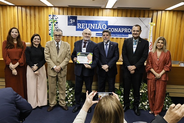 Fotografia de sete pessoas perfiladas lado a lado em p&eacute;, sendo tr&ecirc;s mulheres e quatro homens. Ao centro, um homem exibe um certificado.