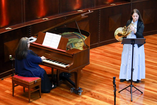 &bull;	&Agrave; esquerda, Clenice Ortilgara est&aacute; sentada a tocar um piano de cauda de madeira, acompanhada por partituras abertas.  &bull;	&Agrave; direita, Lidiane Robaldo. Ela veste uma camisa/vestido longo claro e um casaco/blazer escuro por cima, posicionada atr&aacute;s de um estante de partitura.  &bull;  Cen&aacute;rio: &bull;	O &acirc;ngulo da fotografia &eacute; num plano mais elevado (visto de cima), destacando o ch&atilde;o de madeira polida e a estrutura interna do piano de cauda aberto. 