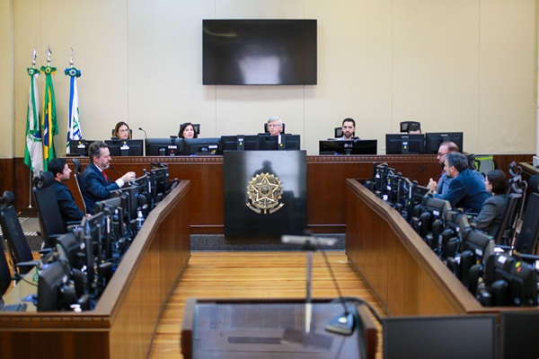 Fotografia de um plen&aacute;rio com mesas e computadores de ambos os lados da imagem, com quatro advogados - dois de cada lado. Ao fundo, mesa de presid&ecirc;ncia da sess&atilde;o, com bras&atilde;o da rep&uacute;blica, e quatro pessoas, doois homens e duas mulheres
