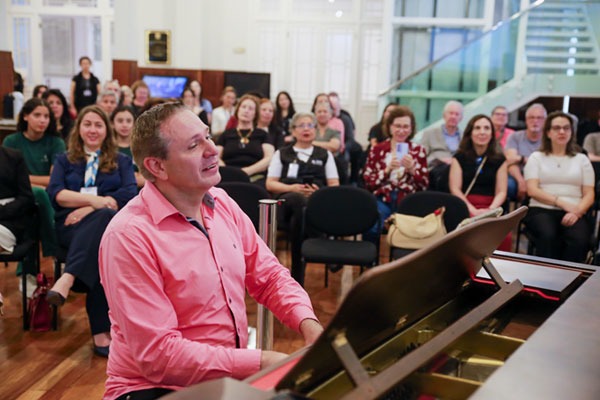 A imagem registra o momento musical durante do evento. Em primeiro plano, o maestro toca piano. Ao fundo, o p&uacute;blico est&aacute; sentado e acompanha a apresenta&ccedil;&atilde;o. Na plateia, pessoas de diferentes idades est&atilde;o acomodadas nas poltronas.