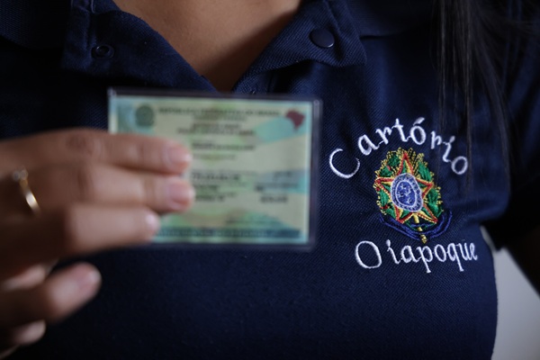 Fotografia de uma mulher segurando um documento de identifica&ccedil;&atilde;o, que est&aacute; desfocado, e com destaquel para a camiseta, escrito: cart&oacute;rio Oiapoque, no entorno do Bras&atilde;o da Rep&uacute;blica