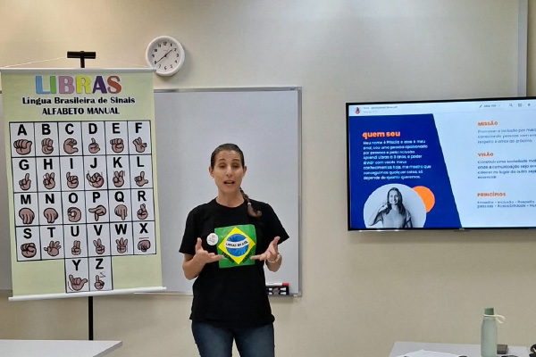 Priscila Luszczynski Barbeta da Silva dando aula de libras Ela usa cabelo apanhado num rabo-de-cavalo, vestida com uma camiseta preta (com a bandeira do Brasil estampada no peito) e cal&ccedil;as de ganga.  &bull;	Est&aacute; de p&eacute;, a falar e a gesticular com as m&atilde;os em frente &agrave; turma.  &bull;  Materiais de Apoio e Apresenta&ccedil;&atilde;o: &bull;	Banner Did&aacute;tico (&agrave; esquerda): Um cartaz num suporte exibe o "ALFABETO MANUAL" da LIBRAS, ilustrando os sinais correspondentes a cada letra de A a Z.  &bull;	Ecr&atilde; de TV/Monitor (&agrave; direita): Um ecr&atilde; fixado na parede mostra um diapositivo de apresenta&ccedil;&atilde;o com as se&ccedil;&otilde;es "Quem sou", "Miss&atilde;o", "Vis&atilde;o" e "Princ&iacute;pios". A sec&ccedil;&atilde;o "Quem sou" apresenta uma breve introdu&ccedil;&atilde;o de uma pessoa chamada Priscila.  &bull;  Espa&ccedil;o e Ambiente: &bull;	Trata-se de uma sala de aula ou audit&oacute;rio bem iluminado, com piso de azulejos claros e paredes brancas.  &bull;	Na parede do fundo, observa-se tamb&eacute;m um rel&oacute;gio anal&oacute;gico e tomadas el&eacute;tricas.  &bull;	Em primeiro plano e nas laterais, v&ecirc;em-se mesas de trabalho brancas, garrafas de &aacute;gua e partes do p&uacute;blico a assistir. 
